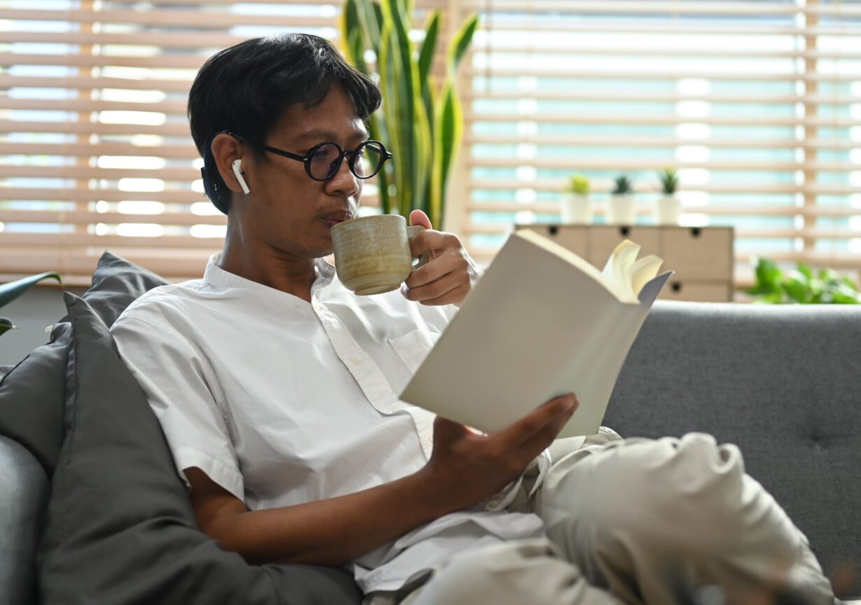 Peaceful asian man drinking hot coffee and reading book on comfortable couch.