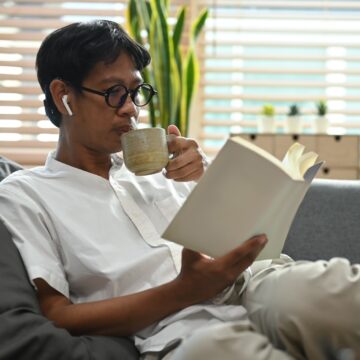 Peaceful asian man drinking hot coffee and reading book on comfortable couch.